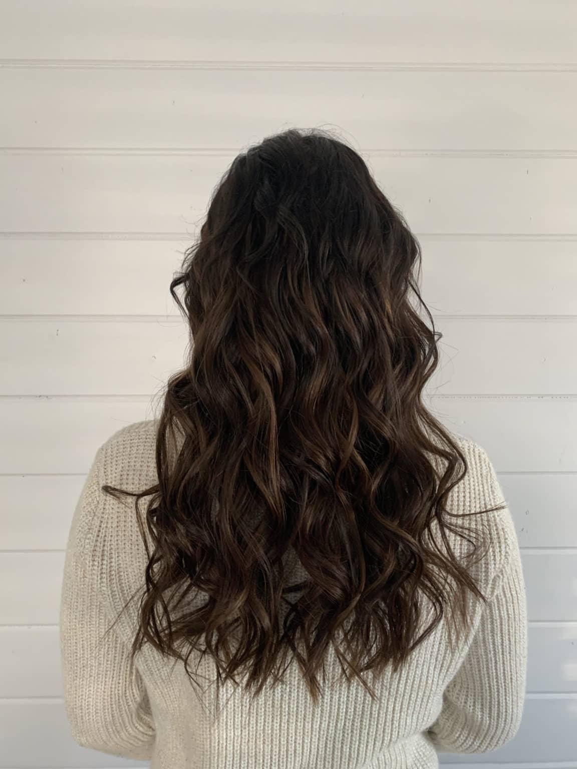Person with long, wavy hair wearing a beige sweater against a light wooden panel background show casing her nano-link hair extensions