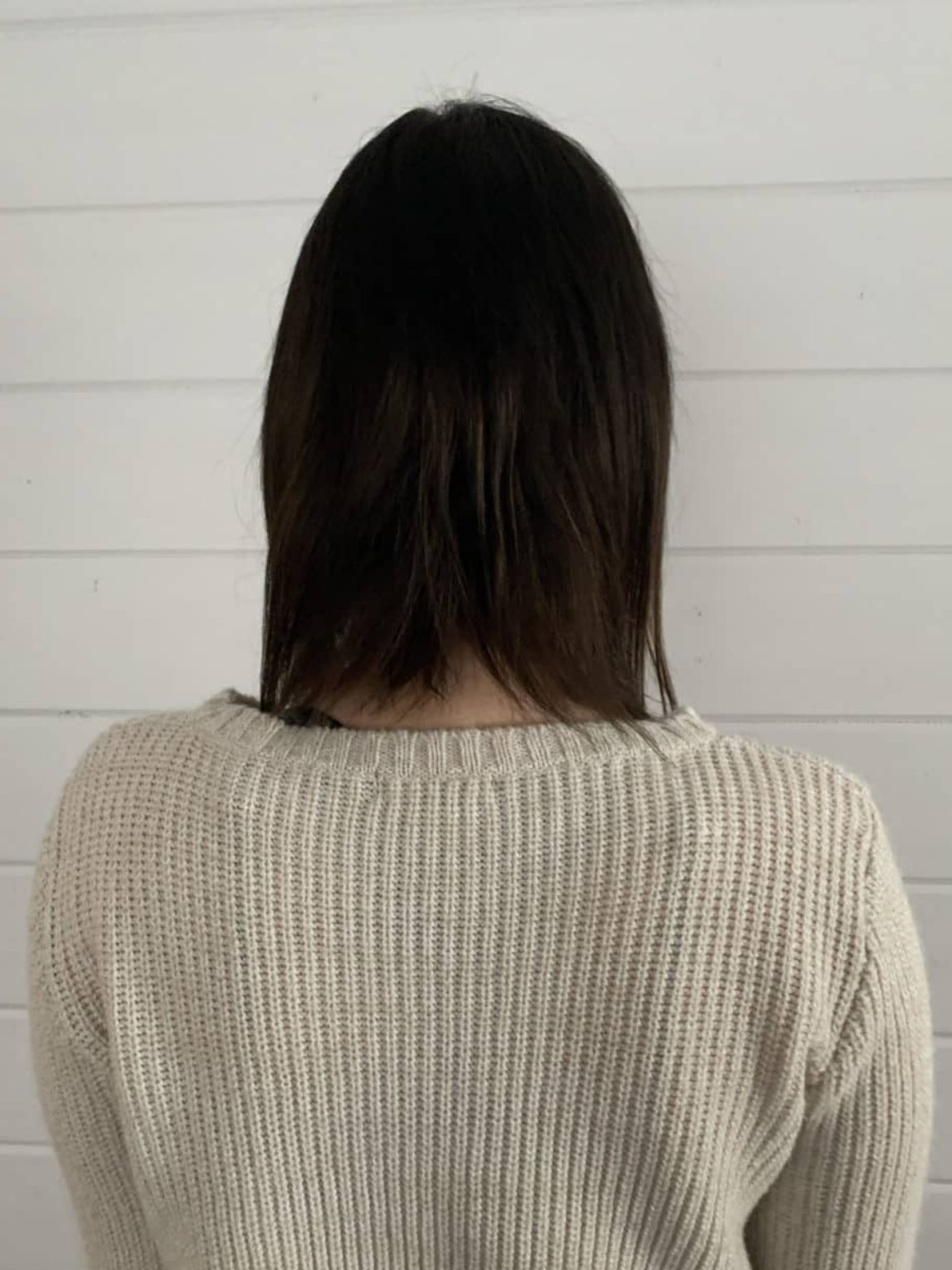 Person wearing a beige sweater against a white wooden panel background showing a before image of her hair